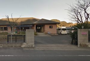 施設の正面外観。ピンク系の建物と駐車場、山を背景とした敷地全体の景観